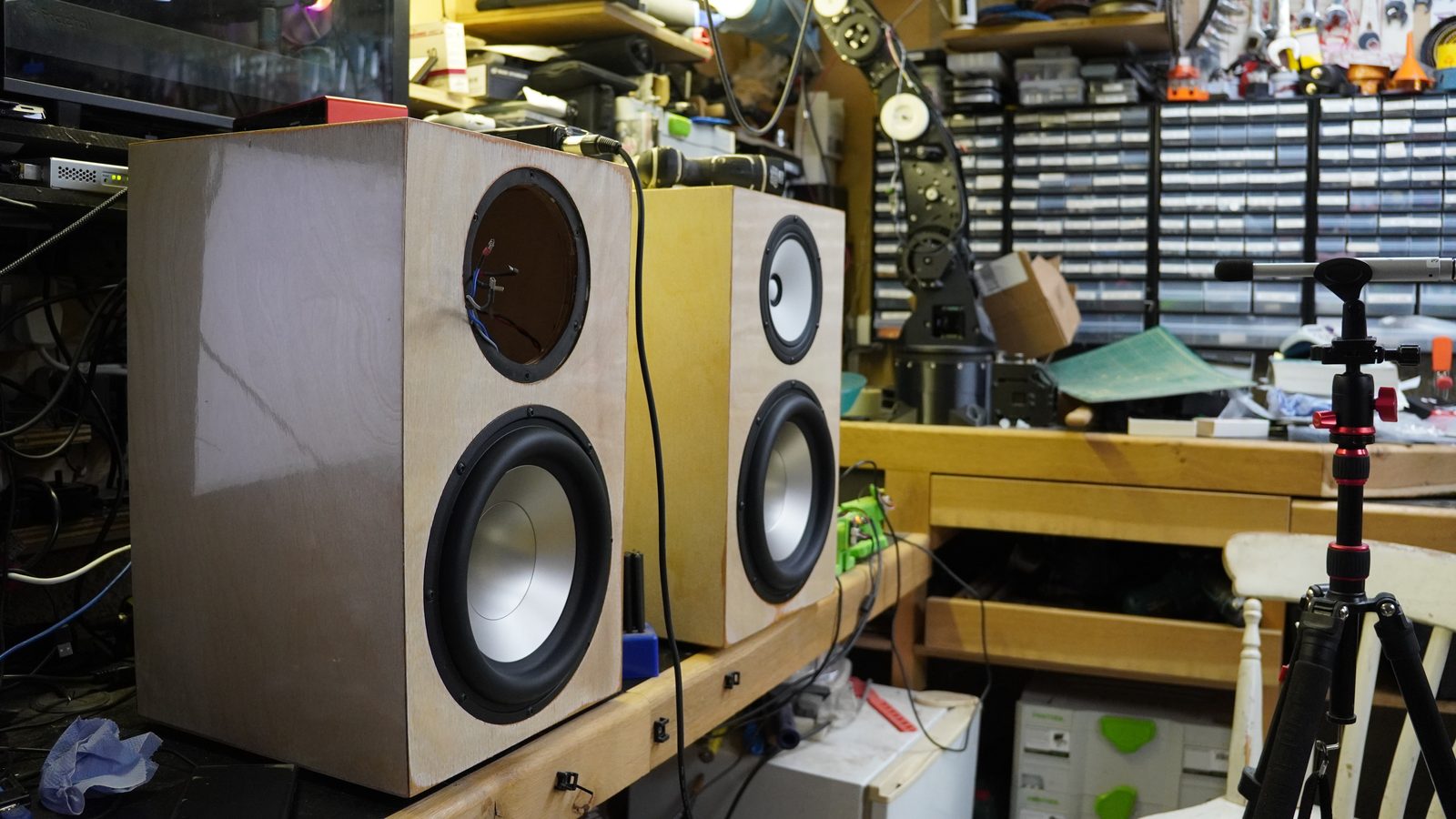 Two unfinished plywood active speakers on a workshop bench during microphone measurement.